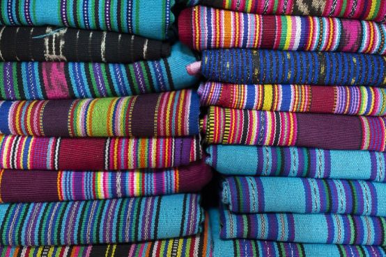 Two piles of brightly coloured woven cloths sit side by side. They are Tais, a traditional fabric from Timor.