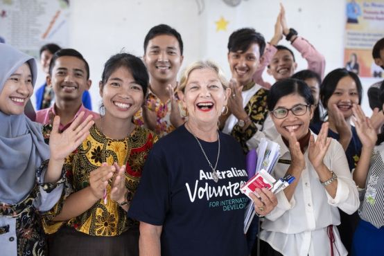 An older lady in an Australian Volunteers t-shirt is surrounded by Indonesian students. They all look very happy and excited.