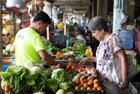 A man in a bright yellow shop shows a lady with grey hair vegetables at a market stall