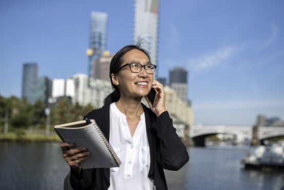 A woman on a mobile phone with a notebook stands in front of a Melbourne skyline