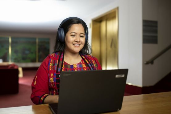 A lady in a red embroidered top sits inside, she is on a laptop and has headphones on