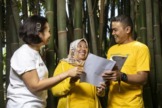 A woman in a white top talks with a woman in a yellow top and head scarf and a man in a yellow top. All look at pieces of paper.