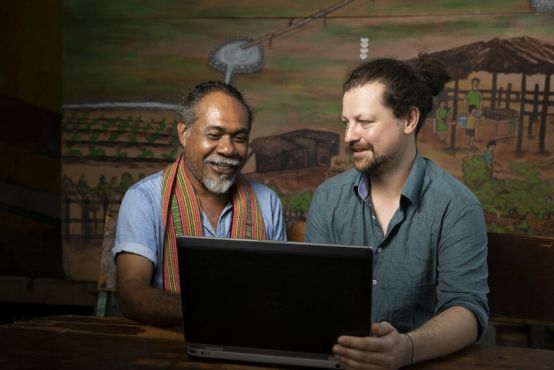 Two men look at a laptop together in front of a painted background of a farm setting