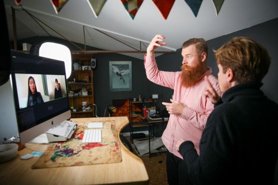 A man with a red beard and red shirt uses sign language. Next to him is a woman in black facing away from the camera. They are looking at a computer screen which has two women on it.