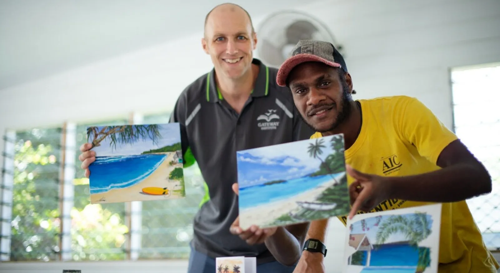 Two people smile into the camera, each holding up a small painting of a realistic coastal scene.
