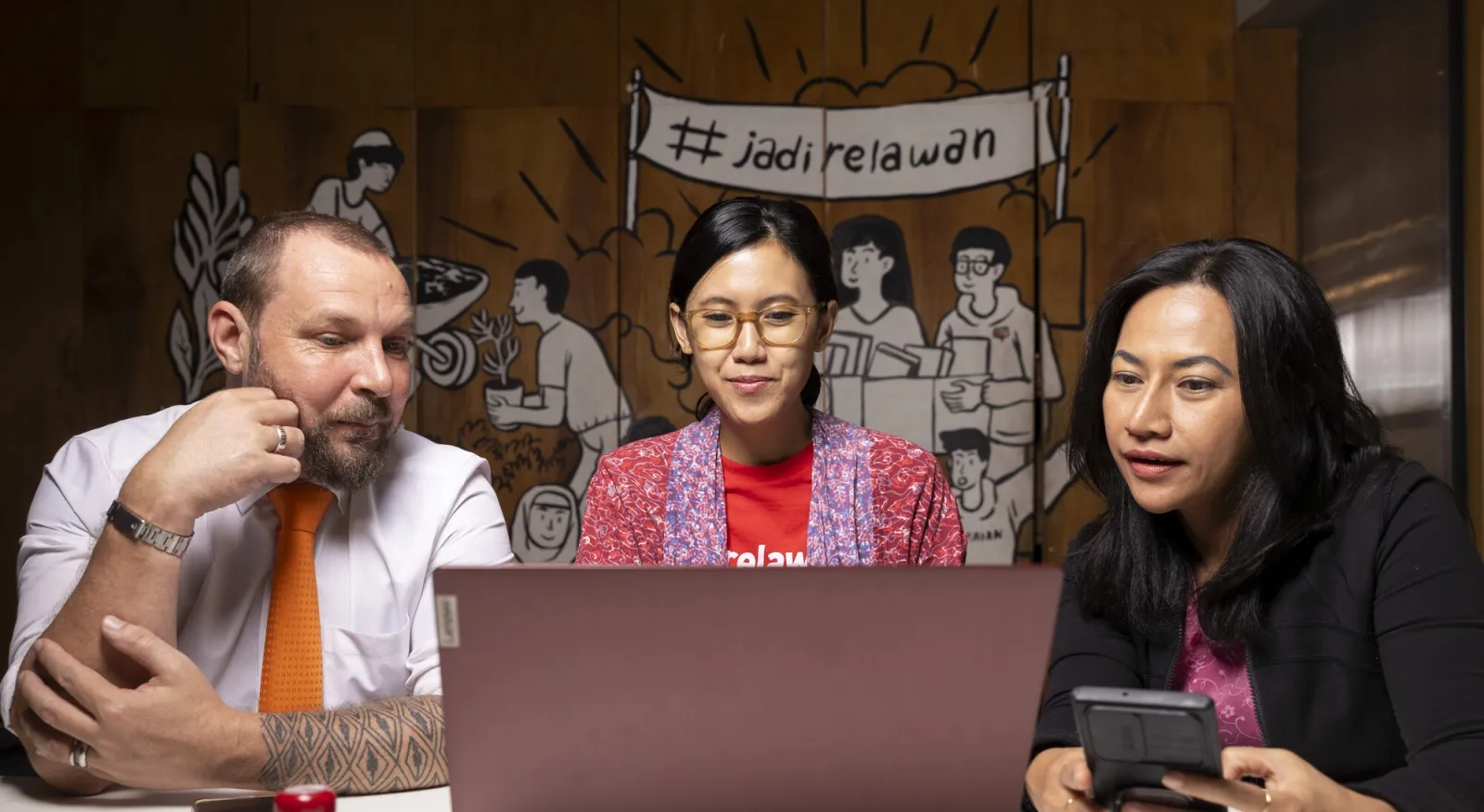 Three people sit in front of a computer, there is a large mural behind them.