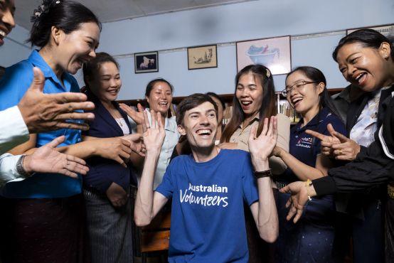 a man in a blue Australian volunteers t-shirt has his hands in the air while his colleagues surround him laughing and clapping. The photo is filled with joy.
